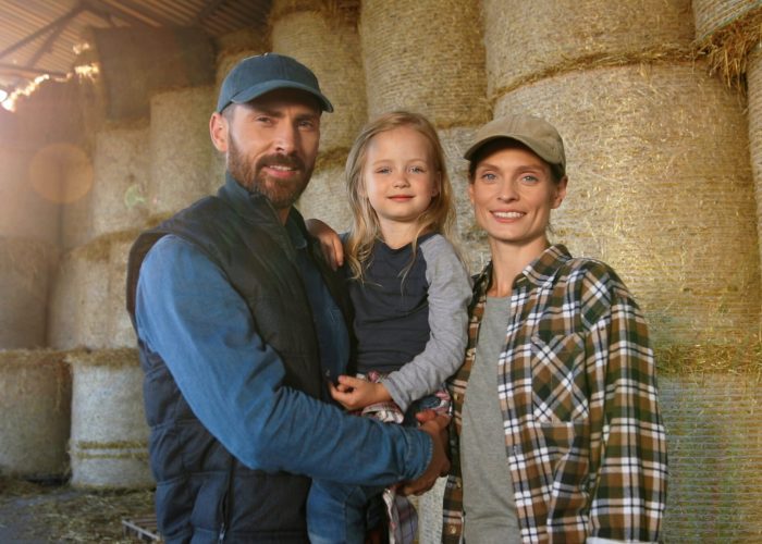 Portrait of happy Caucasian family of farmers with kid standing at barn with hay stocks and smiling to camera. Handsome father, beautiful mother and small pretty daughter at countryside farm.